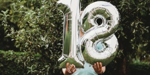 Eine Person steht in einem Garten und hält zwei große Folienballons vor ihren Körper, die eine 18 zeigen.