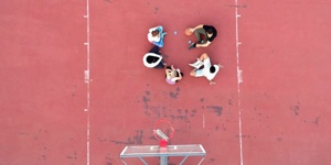 Eine Gruppe von Jugendlichen sitzen auf einem Basketballplatz. Die Fotografie ist aus der Vorgelperspektive aufgenommen.